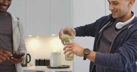 Friends pouring green smoothie in kitchen while using smartphone and enjoying morning