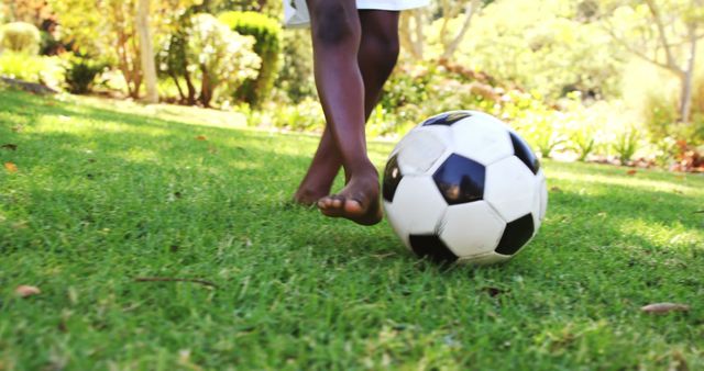 Child seen playing barefoot with a soccer ball in a sunny park. Ideal for promoting outdoor activities, childhood play, summer fun, and children's sports events or soccer. Can be used in advertisements for outdoor equipment, children's sports programs, or summertime family activities.