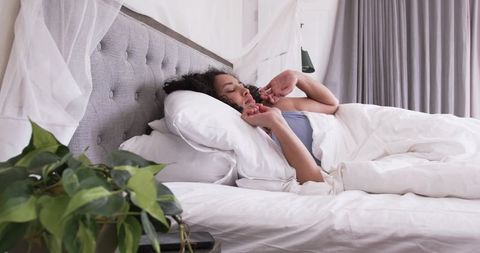 African american woman relaxing in bright bedroom under white duvet with tufted headboard and plant