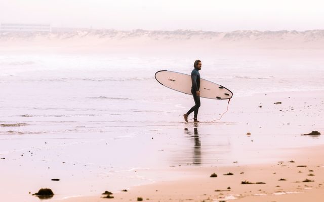 Surfer walking along a sandy beach holding a surfboard during sunset. The calming scene includes ocean waves and the reflection of the surfer on the wet sand. Ideal for themes related to outdoor activities, leisure sport, and beach-side vacations.
