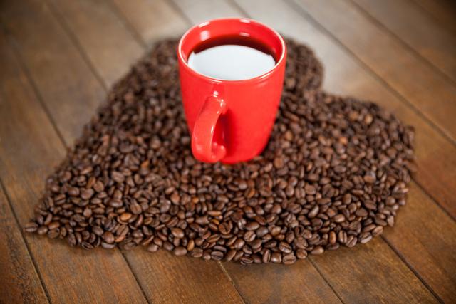 Red coffee mug filled with coffee placed on a heart-shaped arrangement of coffee beans on a wooden surface. This image evokes a sense of warmth and comfort, making it ideal for coffee shop promotions, love-themed advertising, or lifestyle blogs focusing on morning routines and cozy settings.