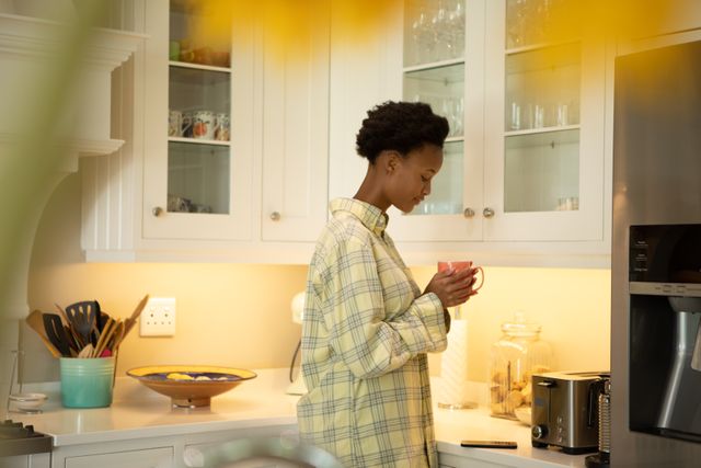 This image depicts an African American woman holding a coffee cup in a modern kitchen, suggesting a relaxed morning routine at home. Ideal for use in lifestyle blogs, home decor websites, and advertisements promoting kitchen appliances or coffee brands.