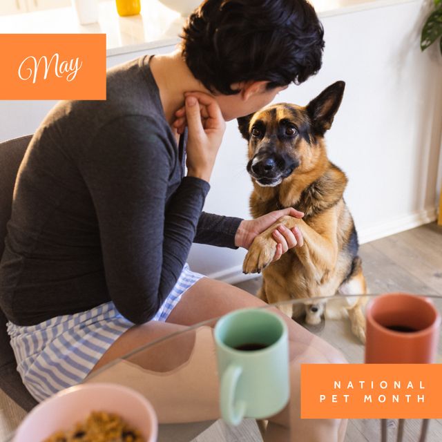 This image depicts a woman holding the paw of a German Shepherd in a home setting, emphasizing the bond between pet and owner during National Pet Month in May. Ideal for promotions related to pet care, grooming, adoption, veterinary services, or any initiative celebrating pets.