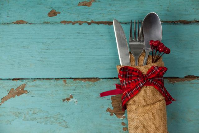 Rustic cutlery wrapped in burlap with a red bow placed on a weathered wooden table. Ideal for use in holiday dining themes, country-style restaurant promotions, or festive event invitations. Perfect for adding a vintage touch to kitchen decor or food-related blog posts.