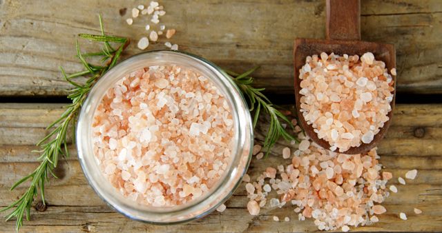 A bowl of pink Himalayan salt is accompanied by fresh rosemary and a wooden scoop on a rustic table, with copy space. Pink Himalayan salt is prized for its purported health benefits and is often used in cooking and as a decorative table salt.