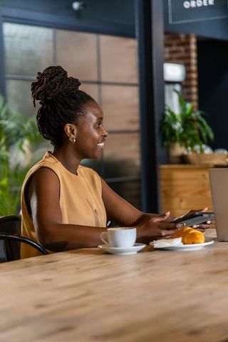 African woman working at cafe with laptop and phone, coffee and croissant on table