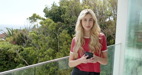 Young blonde woman standing on glass balcony holding smartphone overlooking greenery