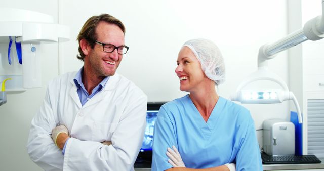 Healthcare professionals, a doctor and a nurse, are smiling and engaged in a friendly conversation in a modern medical clinic. They are both dressed in professional attire: the doctor in a lab coat and the nurse in scrubs and a scrub cap. The background includes typical medical equipment found in a clinic setting, suggesting a high-tech, professional atmosphere. This image can be used to represent teamwork, professional healthcare, medical services, and a positive work environment in healthcare marketing materials.