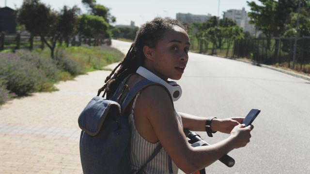 Young African American woman wearing headphones and carrying a backpack, checking her smartphone while standing beside an electric scooter. Ideal for themes related to modern urban transportation, eco-friendly commuting, youthful lifestyle, or technology use in daily life. Great for promoting apps, urban mobility solutions, and outdoor activities.