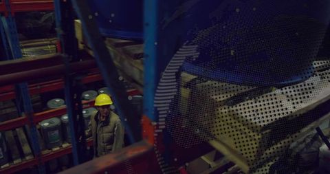 Inspecting warehouse worker wearing yellow hard hat scanning storage racks and drums in dim aisle