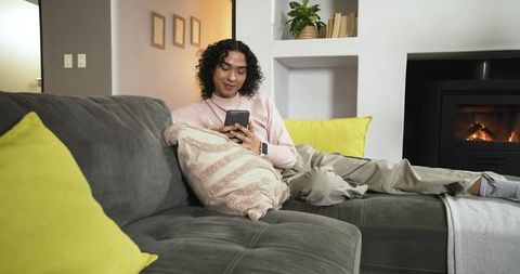 Non-binary person relaxing on gray sectional sofa scrolling smartphone by fireplace