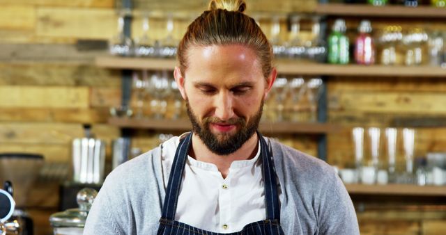 Male barista with beard and man bun stands behind counter, focused on preparing a drink. Kitchen utensils, glassware, and wooden shelves in background create a cozy café atmosphere. Useful for coffee shop advertisements, promotional materials for café equipment, lifestyle blogs, and interior design showcases.