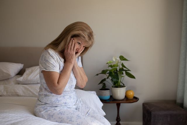 Senior Caucasian woman sitting on her bed with hands on her face, looking tired and sick. Social distancing during Covid 19 Coronavirus quarantine lockdown.
