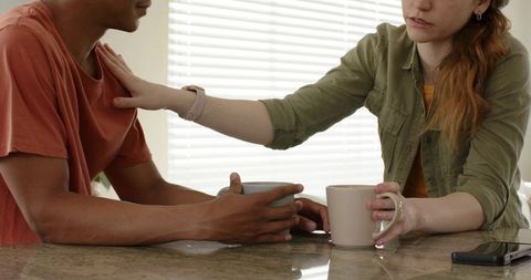 Couple sharing quiet morning at kitchen counter holding mugs and offering supportive touch