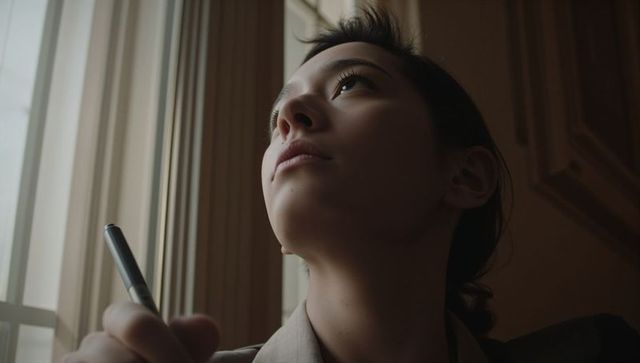 Contemplative asian woman holding pen gazing toward tall window with warm sidelight
