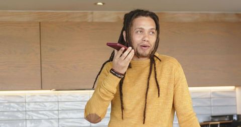 Man speaking into smartphone in modern kitchen wearing mustard sweater and dreadlocks