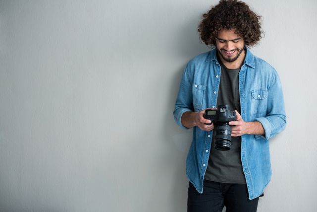 Male photographer with curly hair and casual clothing reviewing captured photos on his digital camera in a studio. Ideal for use in articles about photography, creative professions, modern technology, and hobbies.