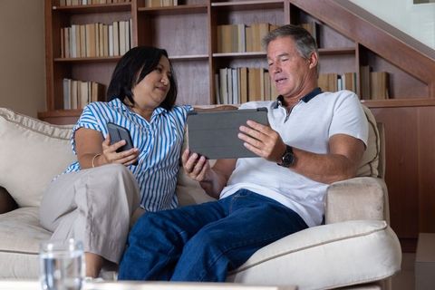 Senior couple relaxing on couch using tablet and smartphone in cozy home library