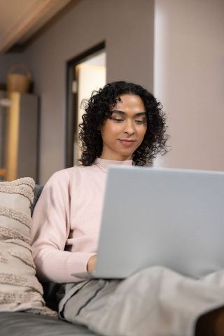 Non-binary person working remotely on laptop, relaxing on couch in pastel pink sweater