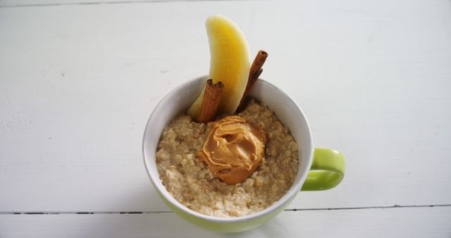A bowl of oatmeal is topped with a dollop of peanut butter, a slice of banana, and a cinnamon stick, with copy space. The image presents a healthy breakfast option, suggesting a nutritious start to the day.