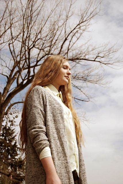 Young woman with long hair standing outdoors on a cloudy day. Bare tree branches stretch out behind her, combining with her light sweater and casual attire to create a tranquil scene. Ideal for use in lifestyle blogs, nature-themed advertisements, or profiles about serenity and relaxation.