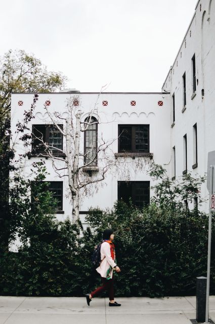 Person is depicted walking past a white historic building with a unique facade, featuring a leafless tree in front. Urban scene embodies mix of vintage architecture and urban greenery. Perfect for use in articles related to city life, historic neighborhoods, urban exploration or residential area imagery.