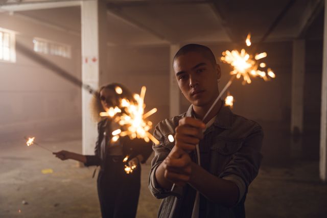 Front view of a hip young biracial man and a hip young Caucasian woman in an empty warehouse, holding sparklers, looking straight to the camera.