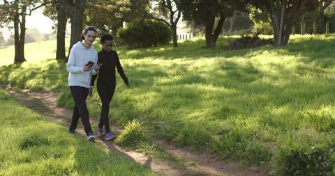 Young diverse friends walking along sunlit meadow trail checking phone casual stroll