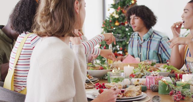Group of friends celebrate Christmas around decorated table filled with festive food. Christmas tree and gifts in background add to holiday atmosphere. Image ideal for holiday greetings, social media posts about Christmas, and festive season advertising.