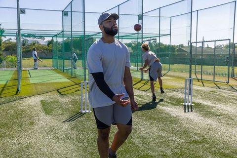 Men practicing cricket in outdoor nets bowling and fielding with red ball on turf