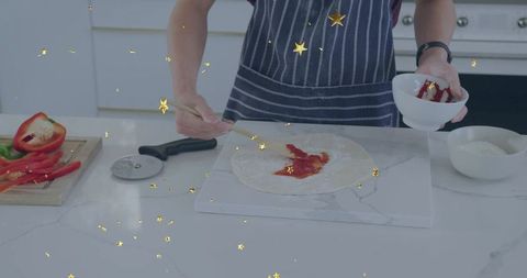 Teen spreading tomato sauce on pizza dough wearing navy striped apron in home kitchen