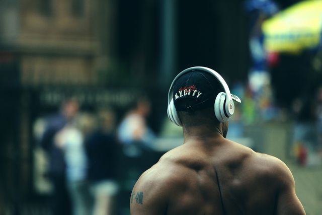 A muscular man wearing wireless headphones is standing outdoors. He is seen from the back, highlighting a tattoo on his shoulder and his muscular physique. This can be used for depicting modern fitness lifestyle, relaxation while listening to music, or technology in daily life.