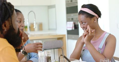 Teen wearing pink headband and denim overalls talking and gesturing with family at kitchen table