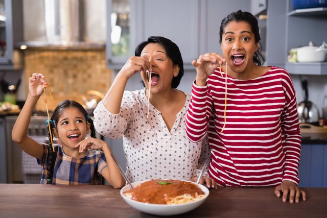 Three generations of women enjoying a meal together in a home kitchen. They are smiling and playfully eating spaghetti, creating a joyful and warm atmosphere. This image is ideal for illustrating family bonding, home-cooked meals, and the joy of spending time with loved ones. Perfect for use in advertisements, blogs, and articles related to family life, cooking, and home dining.