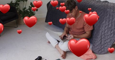 Young man using laptop and phone on bedroom floor with floating heart icons
