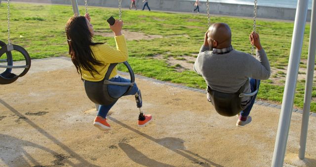 Two friends with prosthetic legs are enjoying a swing set in a park on a sunny day. This image can be used to promote inclusivity, outdoor recreation, and friendship. It highlights the fun and joy irrespective of physical challenges, perfect for campaigns and publications focused on disability awareness, community activities, and adaptive sports.