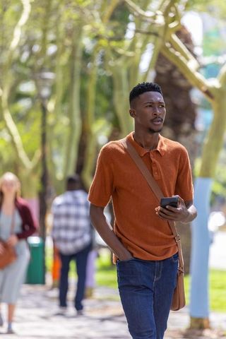 African american man walking holding phone wearing rust polo and crossbody bag under tree canopy