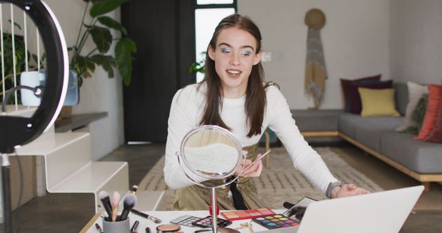 Young woman enthusiastically filming makeup tutorial with various cosmetics in front of her, surrounded by makeup tools and using a ring light and mirror. Ideal for illustrating beauty vlogging, makeup tutorials, influencer marketing, at-home filming setups, and social media content creation.
