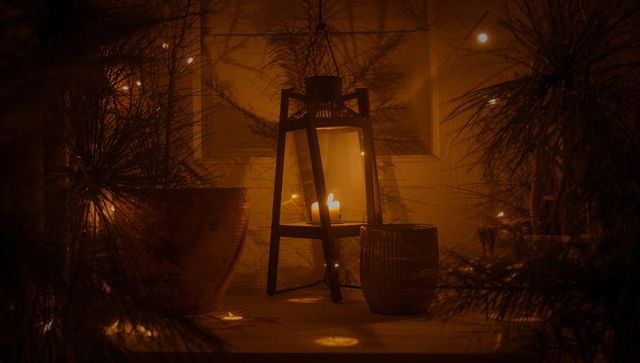 Black-framed lantern casting warm amber glow on window ledge with candles and pine bokeh