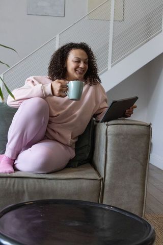 African american woman relaxing on sofa with mug and tablet in pale pink loungewear