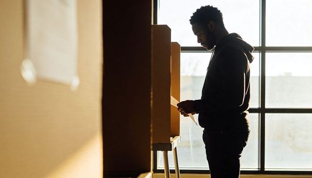 Backlit voter casting ballot at cardboard privacy booth by sunlit window
