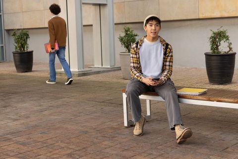 Diverse students relaxing on campus bench using smartphone with notebooks and binders