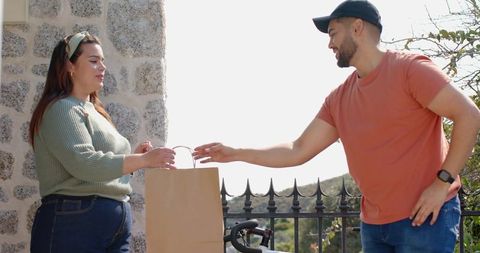 Courier handing brown paper bag to neighbor on stone porch beside bicycle and gate