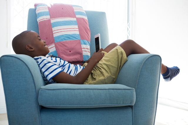 Side view of smiling boy looking at mobile phone while lying on armchair in sitting room