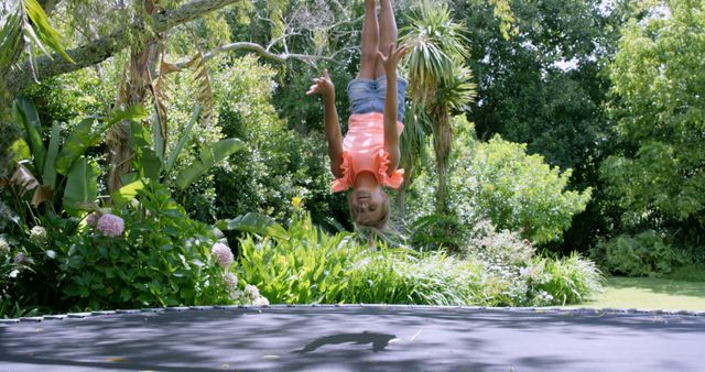 A young Caucasian girl is enjoying herself while upside down in mid-air on a trampoline, surrounded by lush greenery, with copy space. Her playful activity captures the essence of carefree childhood moments outdoors.