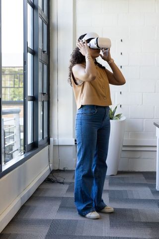 African american woman adjusting vr headset in modern sunlit office wearing denim jeans