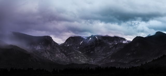 Panoramic view of misty mountains shrouded in dark, stormy clouds. The majestic peaks surrounded by dense fog present a dramatic and moody atmosphere. Ideal for use in travel brochures, nature documentaries, or as a backdrop for presentations on environmental topics.