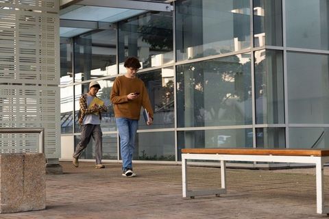 Male students walking beside glass campus building checking smartphone carrying folders