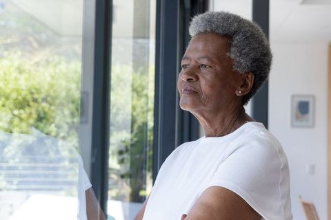 Senior african american woman standing at window looking contemplative toward sunlit garden