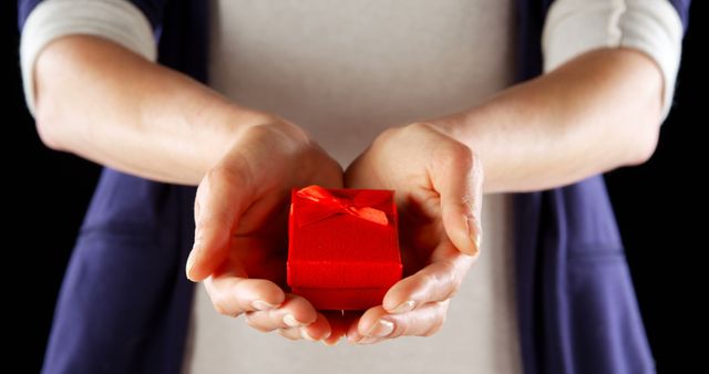 A Caucasian woman presents a small red gift box with a ribbon, symbolizing a gesture of giving or surprise, with copy space. Her hands are gently cupped around the box, emphasizing the care and thoughtfulness behind the gift.